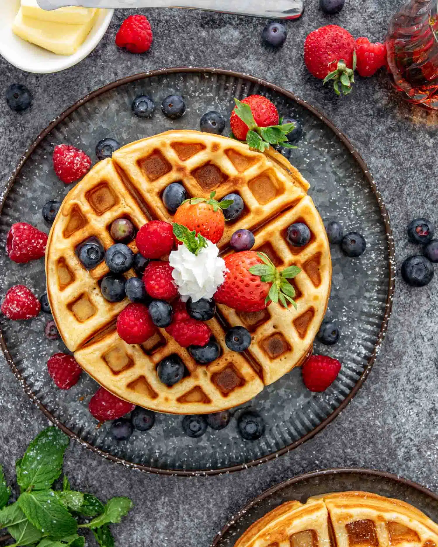 a stack of waffles on a metal plate surrounded topped with berries and whipped cream.