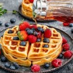 2 large round waffles on a plate topped with maple syrup, whipped cream and berries.