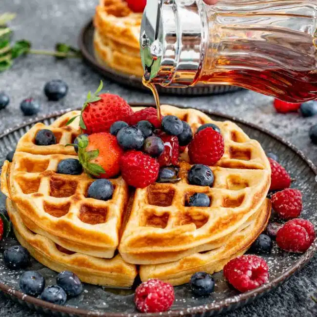 2 large round waffles on a plate topped with maple syrup, whipped cream and berries.