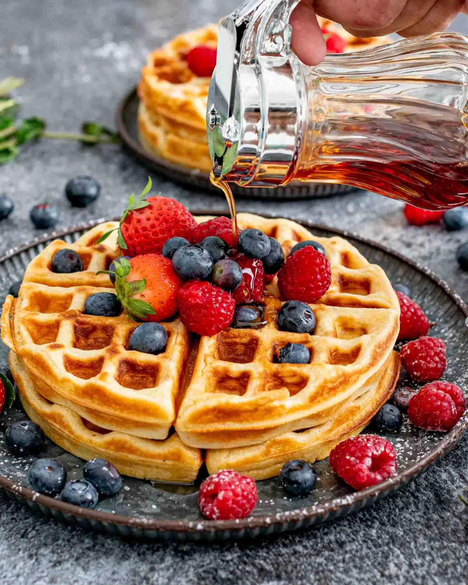 2 large round waffles on a plate topped with maple syrup, whipped cream and berries.