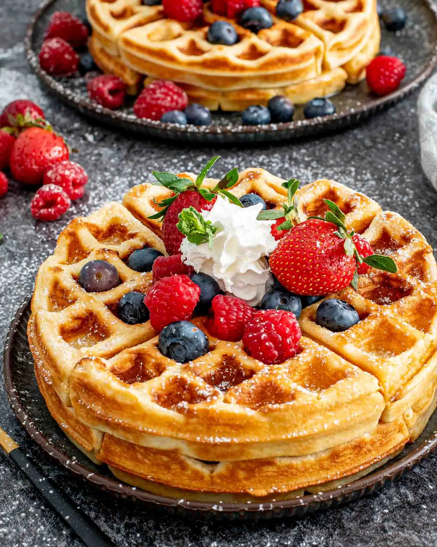 2 large round waffles on a plate topped with maple syrup, whipped cream and berries.