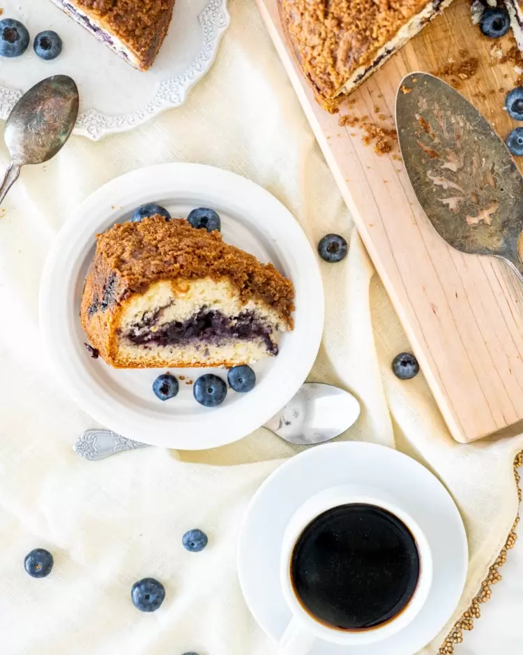 overhead shot of a slice of blueberry coffee cake on a white plate and a cup of coffee