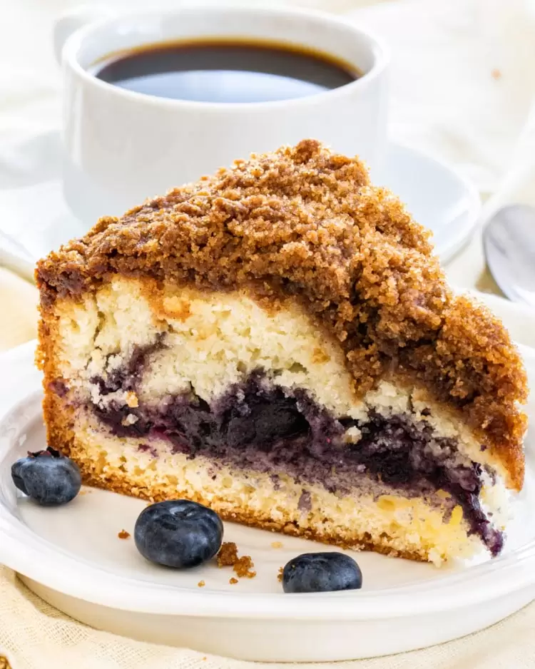 a slice of blueberry coffee cake on a white plate with a cup of coffee in the background