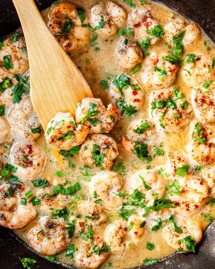 overhead shot of shrimp scampi in a skillet with a wooden spoon holding 3 shrimp
