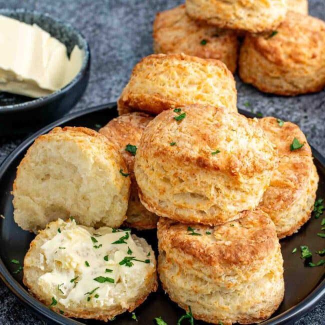 a few buttermilk parmesan biscuits on a black plate with one cut in half and spread with butter.
