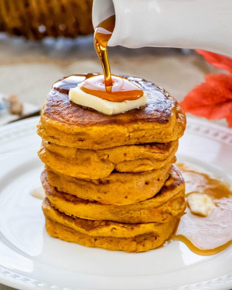 pouring maple syrup over a stack of pumpkin pancakes