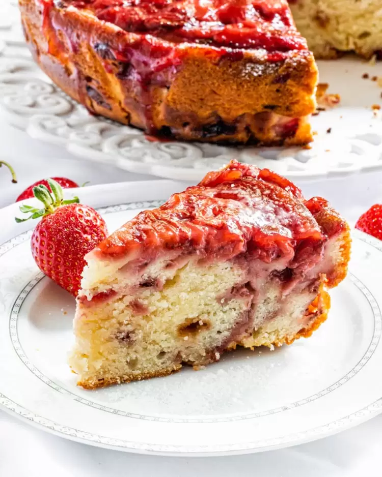 a slice of strawberry cake on a white plate with the whole cake in the background on a platter