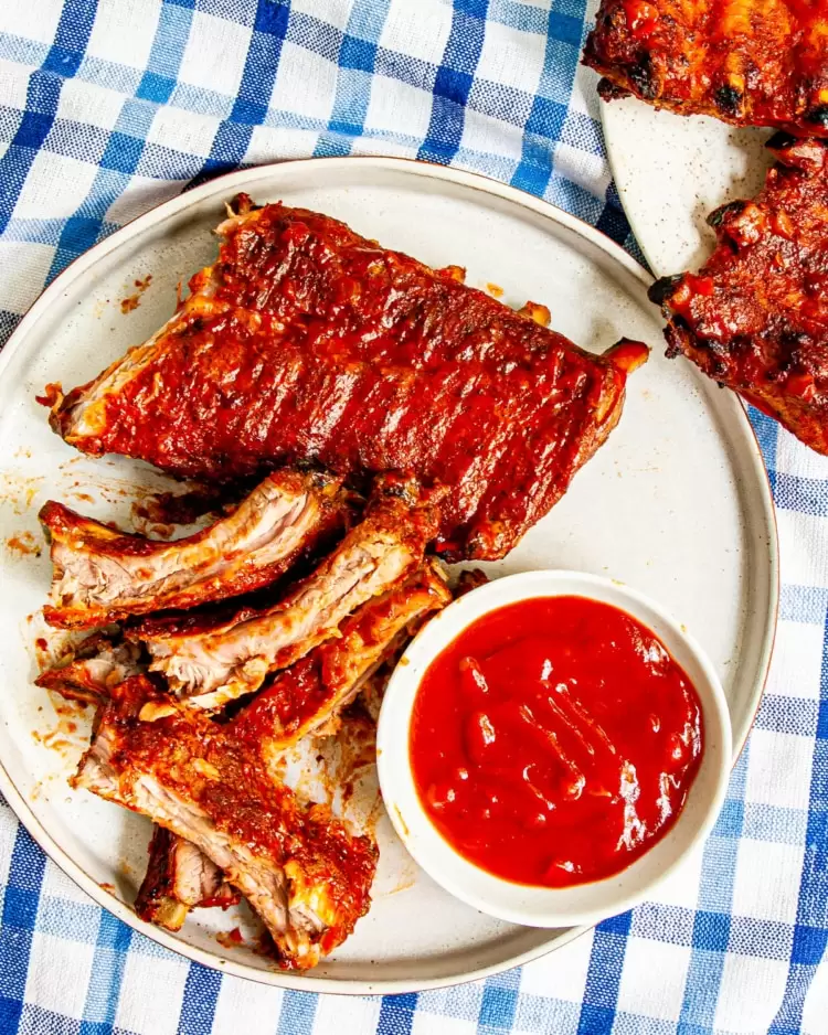 overhead shot of a rack of bbq baby back ribs cut in pieces with more bbq sauce in a white bowl on white plate