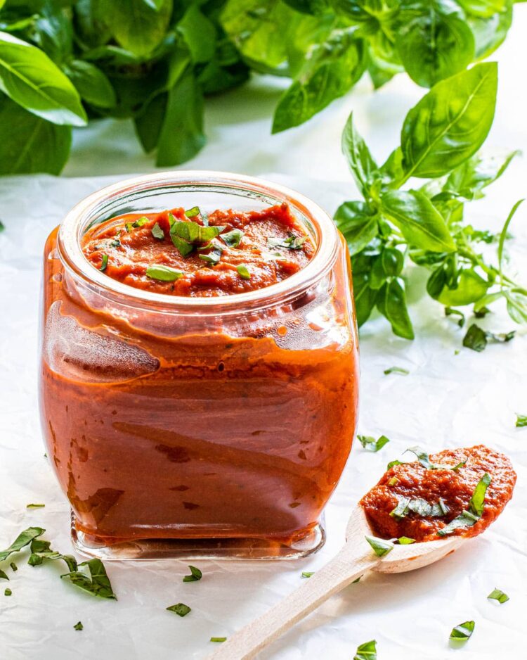a jar of marinara sauce with basil in the background and a spoon next to it