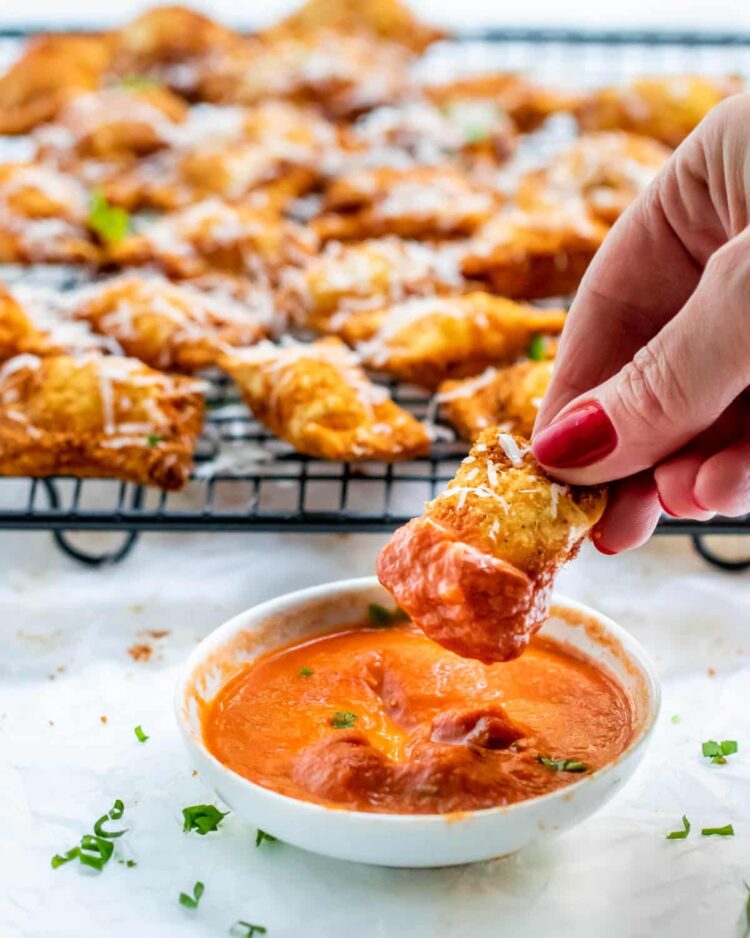 a hand dipping a fried ravioli in a little bowl with marinara sauce