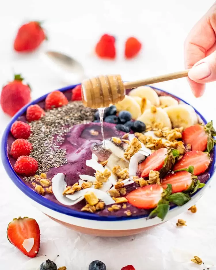 an açai bowl topped with bananas and berries with a hand drizzling honey over it