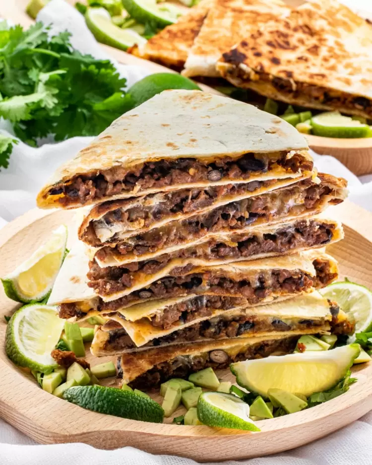 a stack of cheese beef quesadillas on a wooden plate garnished with limes and avocados