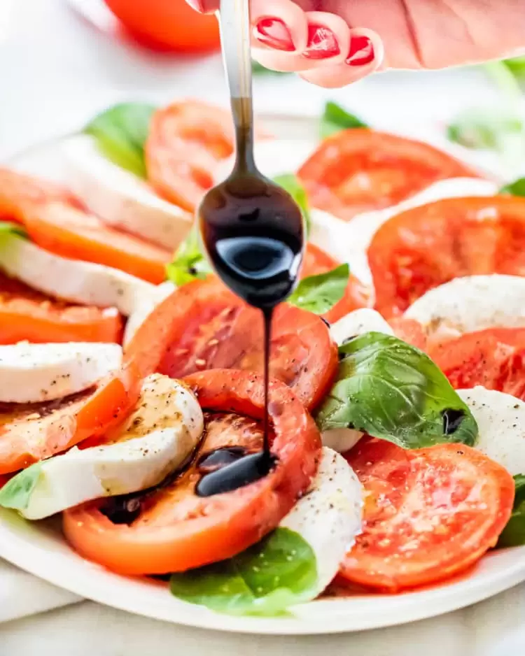 a hand holding a spoon drizzling balsamic glaze over caprese salad