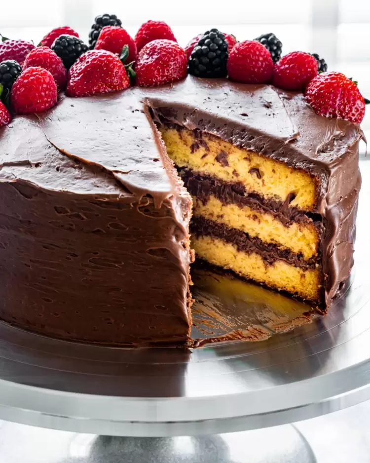 yellow cake with chocolate frosting decorated with berries, with a slice cut out of it