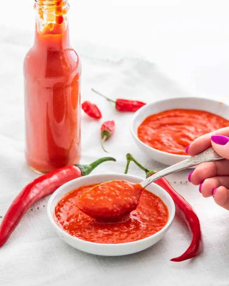 a hand holding a tsp of hot sauce over a bowl filled with hot sauce