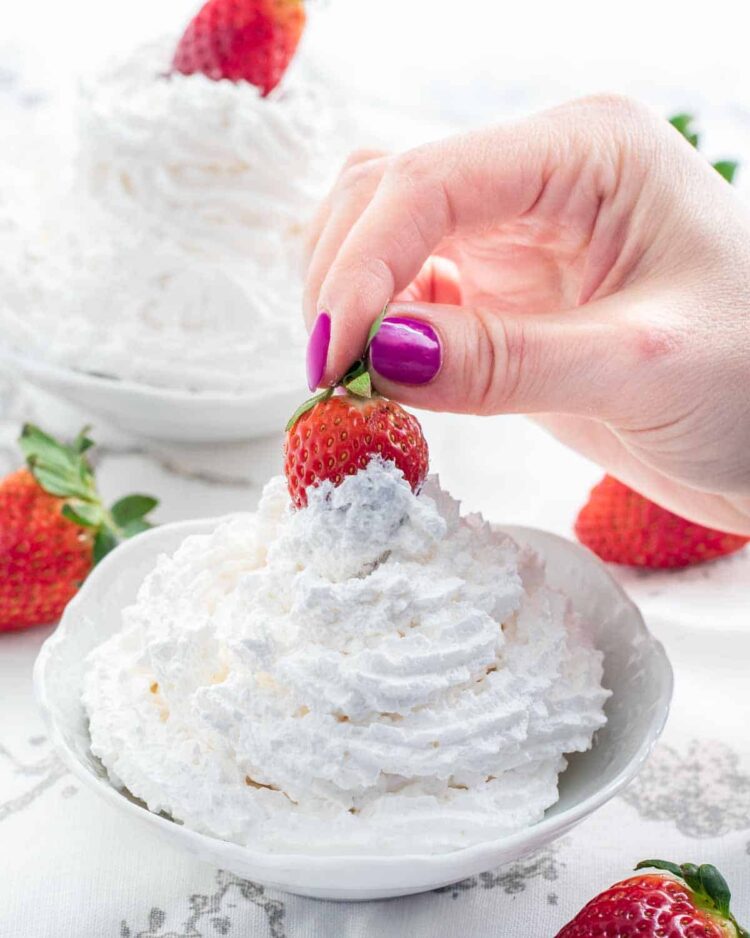 a hand dipping a strawberry in a bowl filled with whipped cream