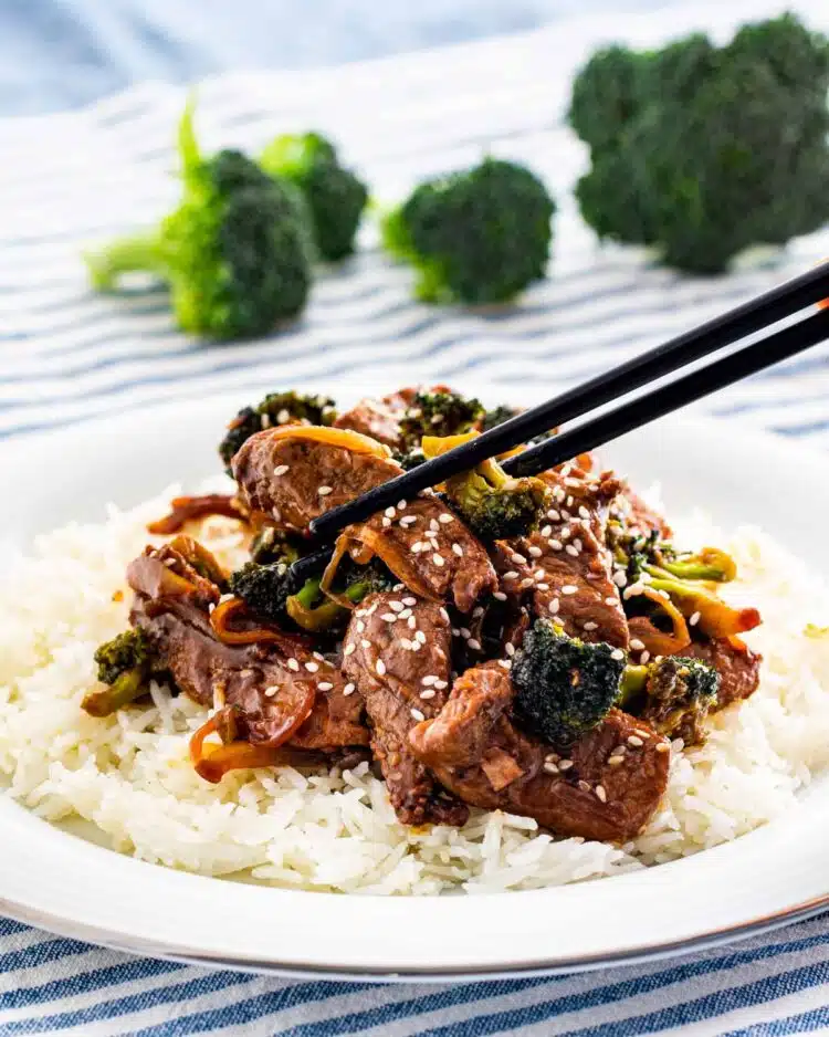 beef and broccoli over a bed of rice garnished with sesame seeds and a pair of chopsticks picking up a piece of meat