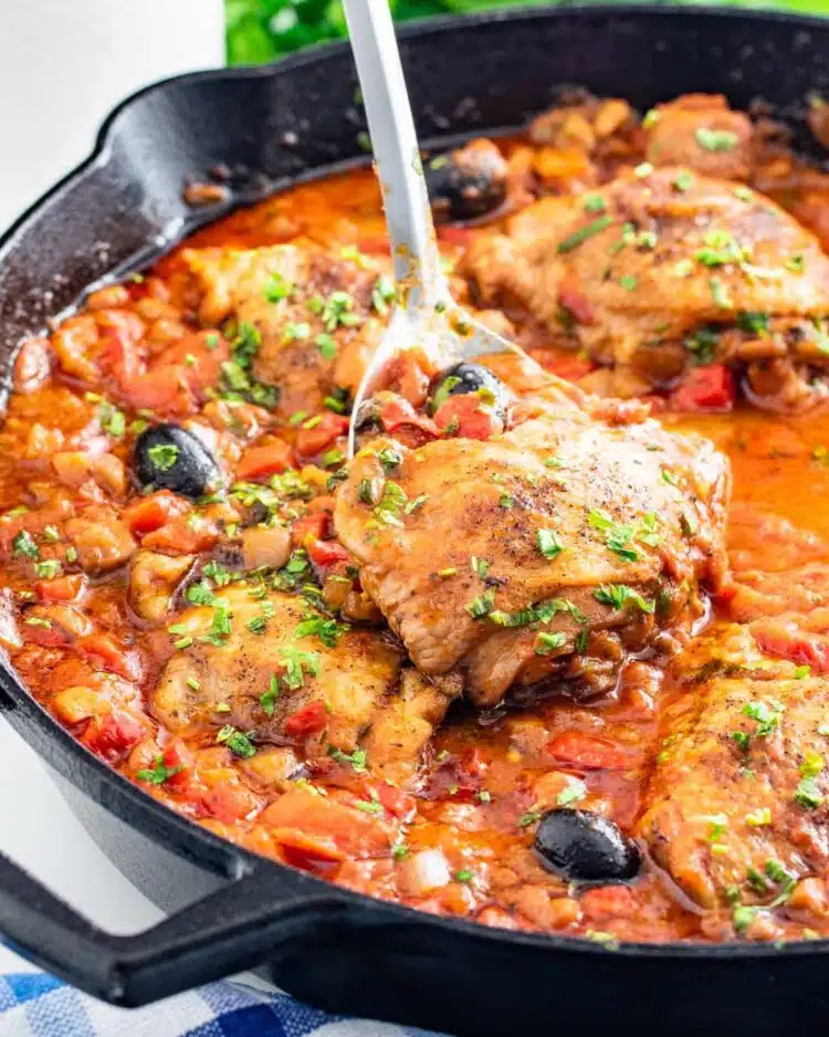 side shot of freshly made chicken cacciatore in a black skillet with a serving spoon inside lifting a chicken thigh