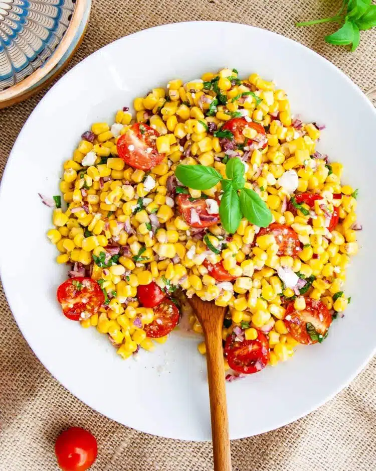 overhead shot of corn salad in a white plate with a wooden spoon inside and a basil leaf on top