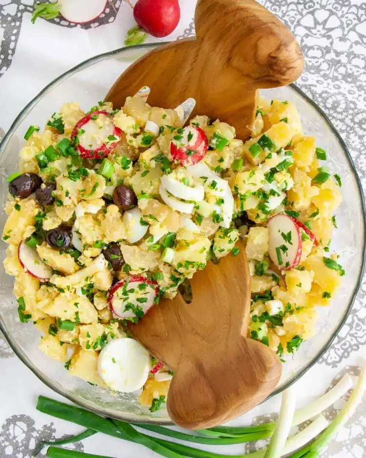 overhead shot of spring potato salad in a glass bowl with two tossing salad hands