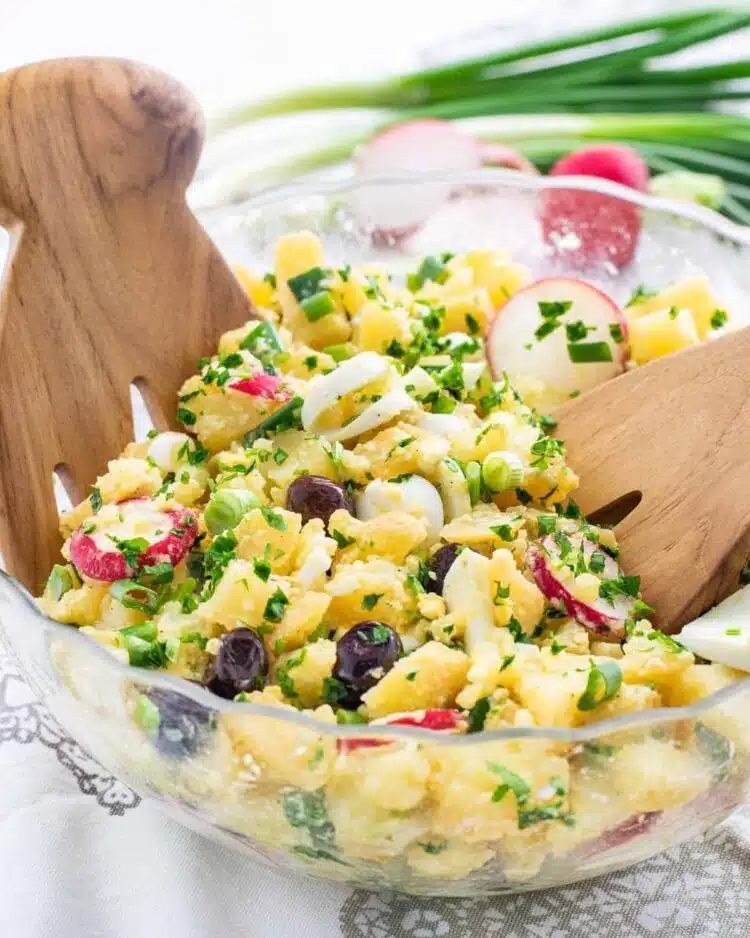 a glass bowl full of spring potato salad with two tossing salad hands in it