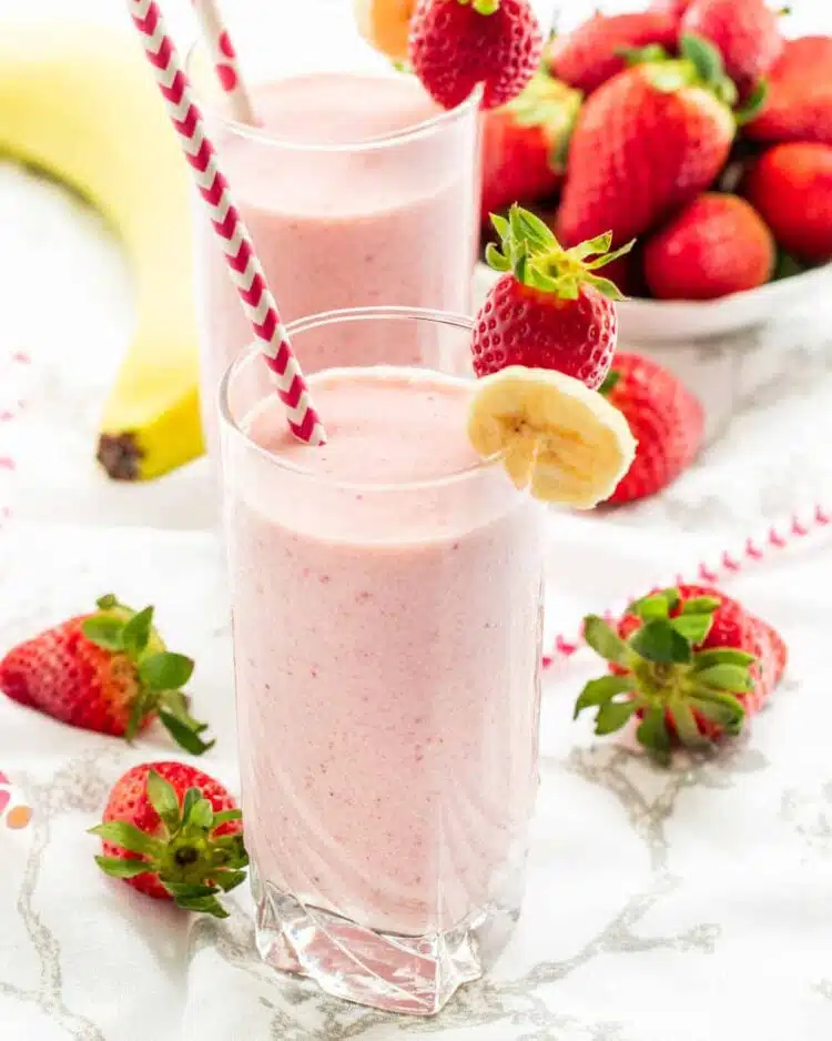 two glasses filled with strawberry banana smoothie  surrounded by strawberries