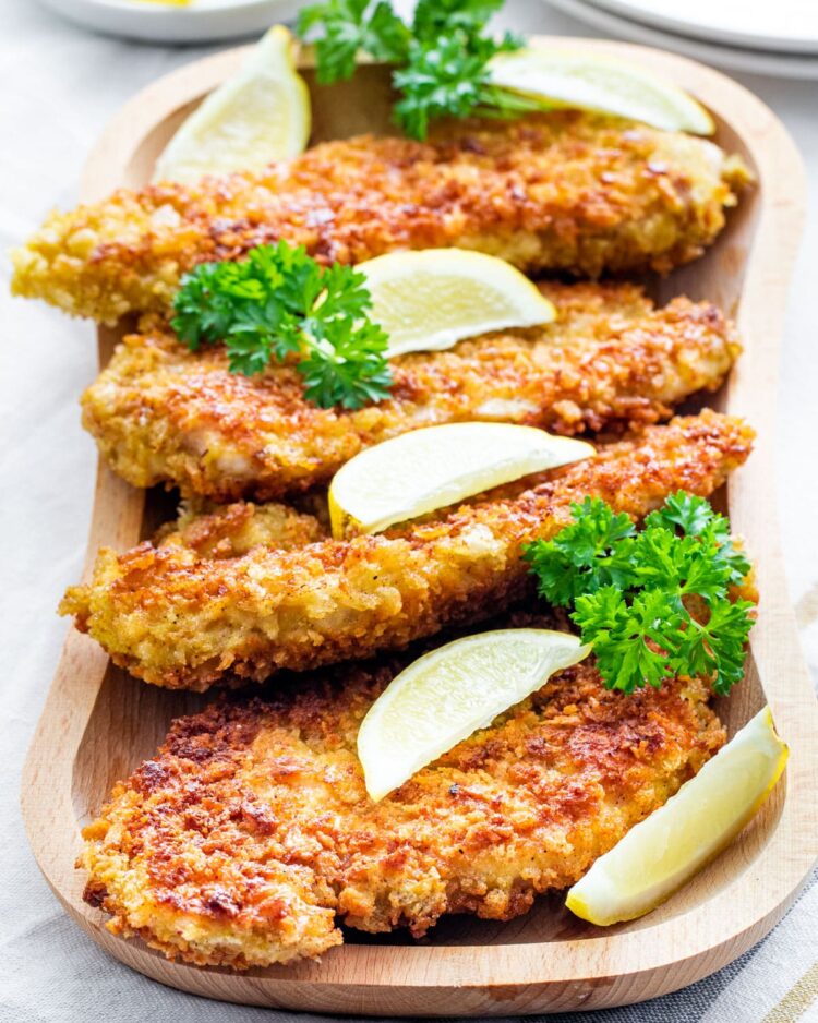 sideview shot of chicken cutlets on a wooden platter garnished with lemon wedges and parsley