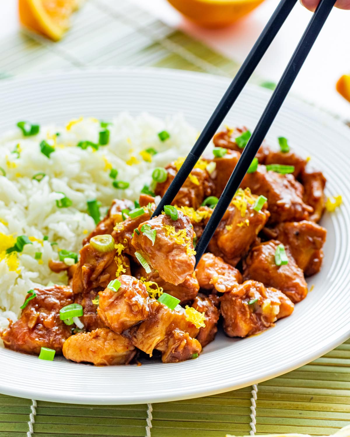 orange chicken on a white plate next to a bed of rice garnished with green onions and lemon zest