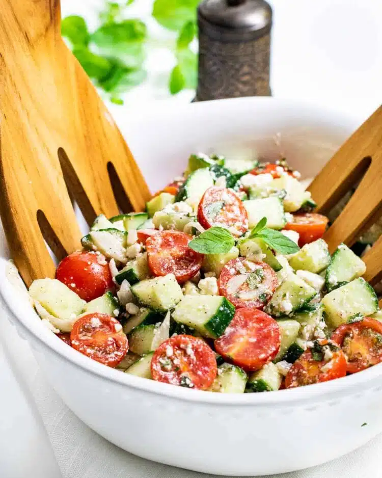 side view shot of cucumber tomato salad in a white bowl with two salad hands in it