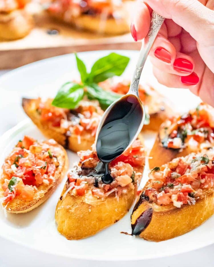 a hand drizzling a tsp of balsamic reduction over bruschetta on baguette slices