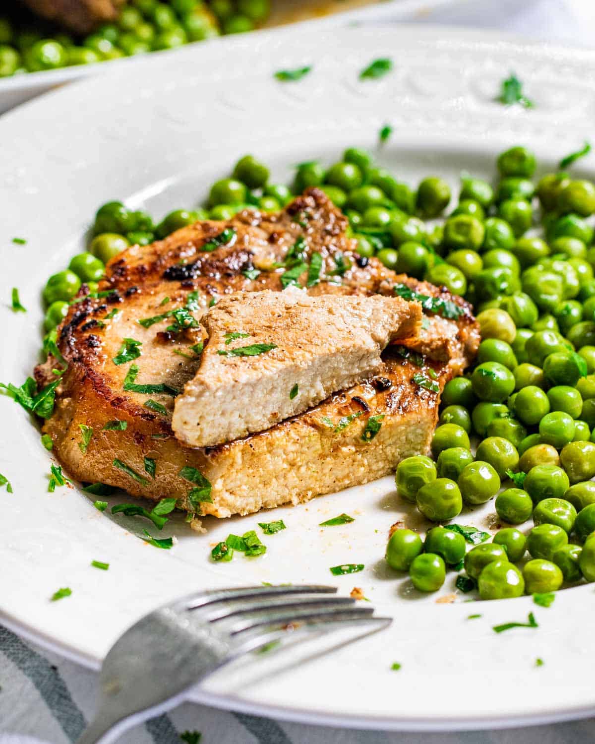 side view shot of a lemon garlic pork chop cut in half and stacked with a side of peas on a white plate