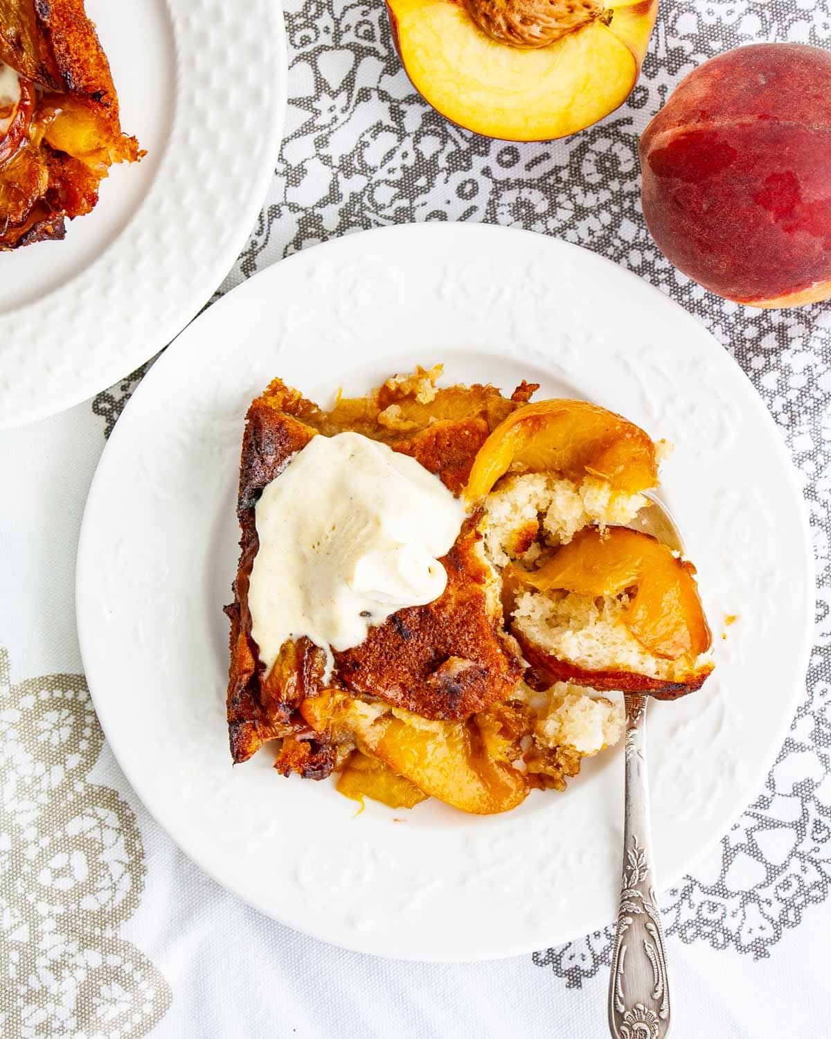 overhead shot of a piece of peach cobbler with a scoop of vanilla ice cream on a white plate with a spoon 