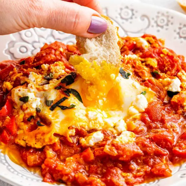a hand dipping a piece of bread in a plate with shakshuka.
