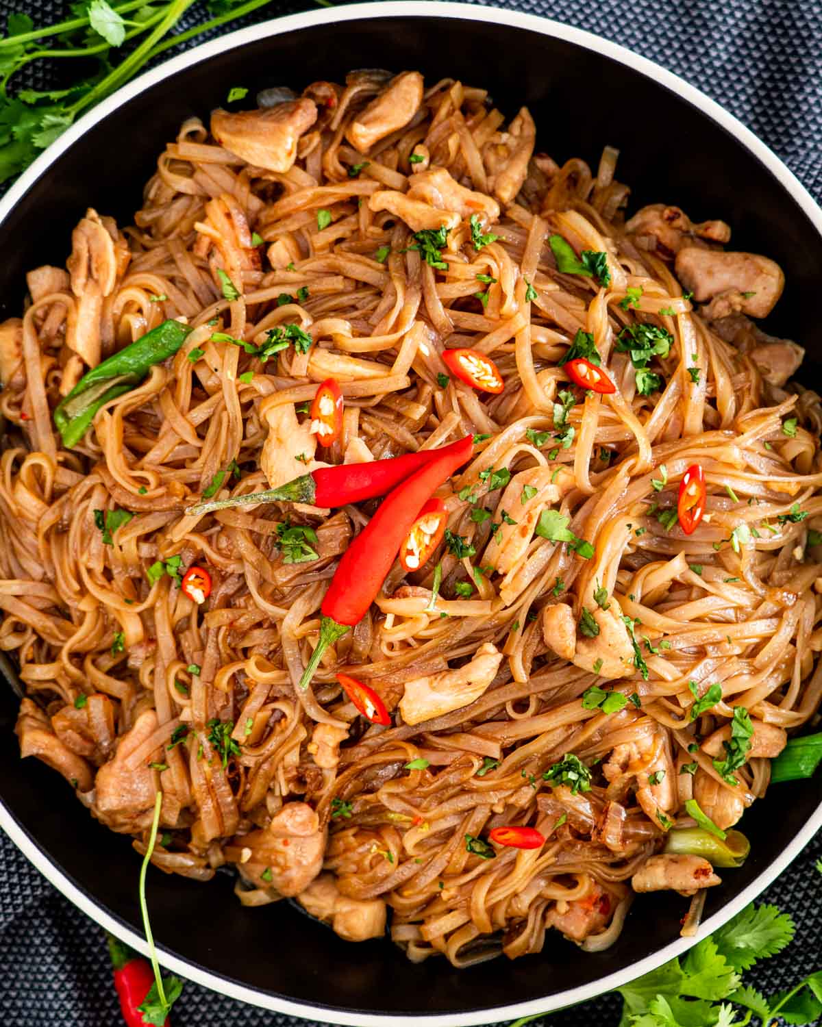 thai drunken noodles in a black bowl garnished with 2 red chilies.