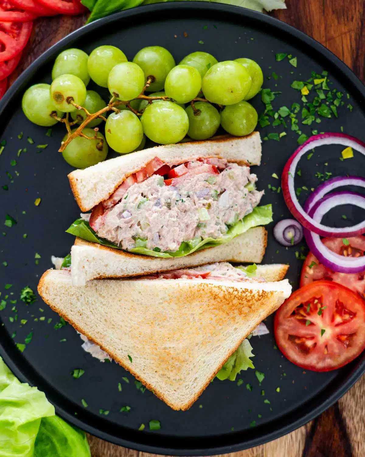 tuna salad sandwich with lettuce and tomatoes and grapes on the side on a black plate.