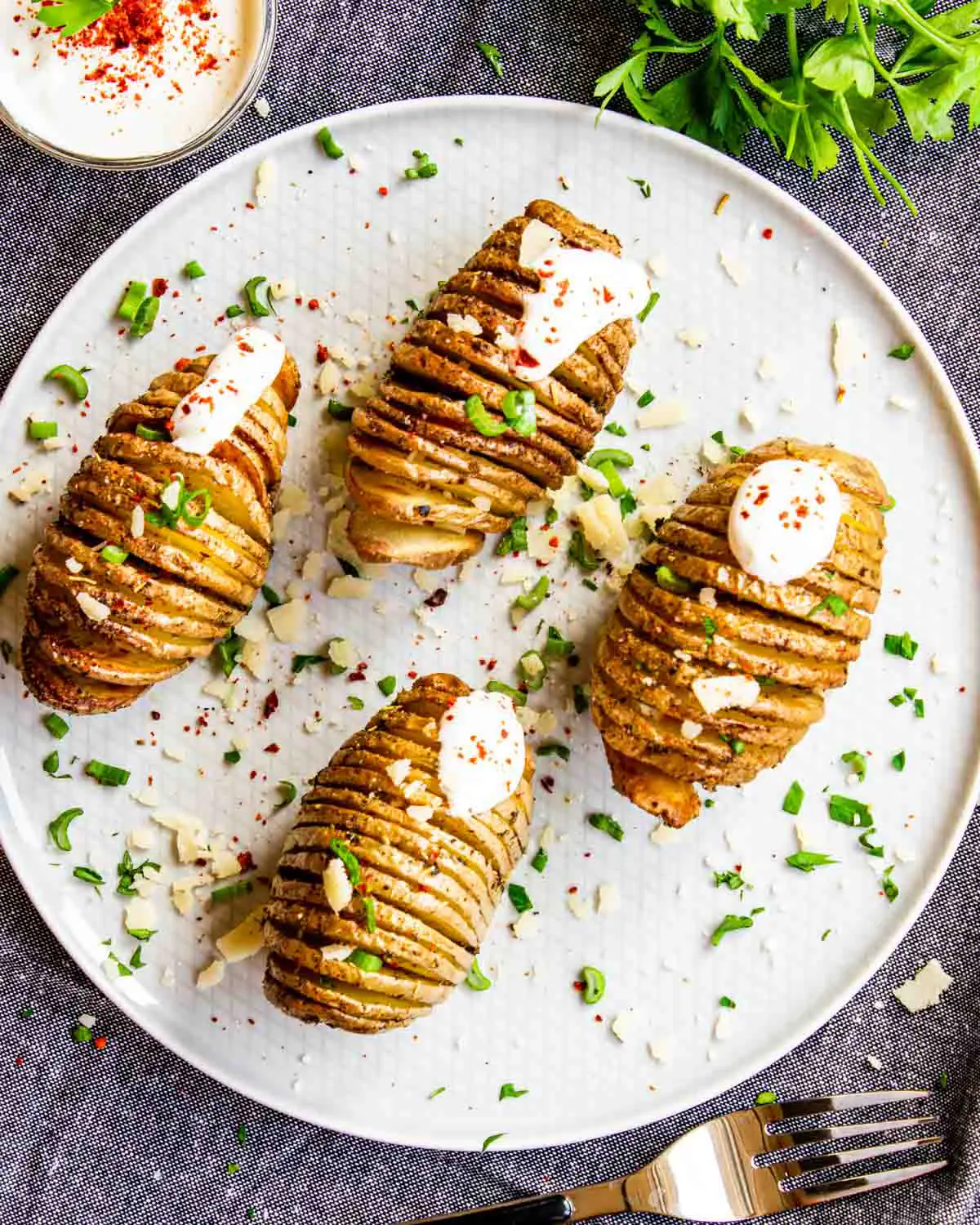 4 hasselback potatoes on a white plate with sour cream and parsley.