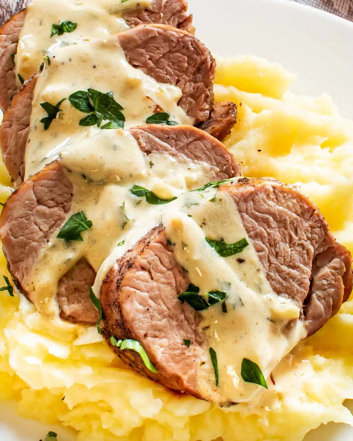 sliced up pork tenderloin with mustard sauce over a bed of mashed potatoes garnished with parsley.