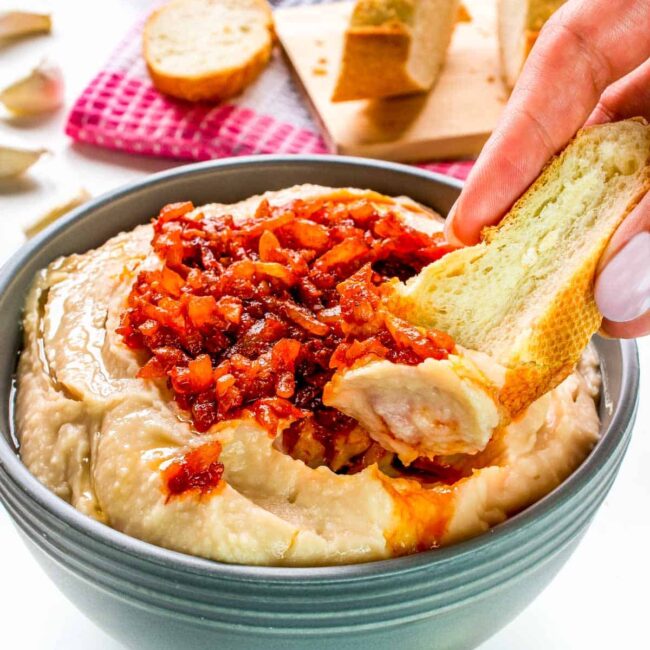 a hand dipping a piece of bread into a bowl with white bean dip.