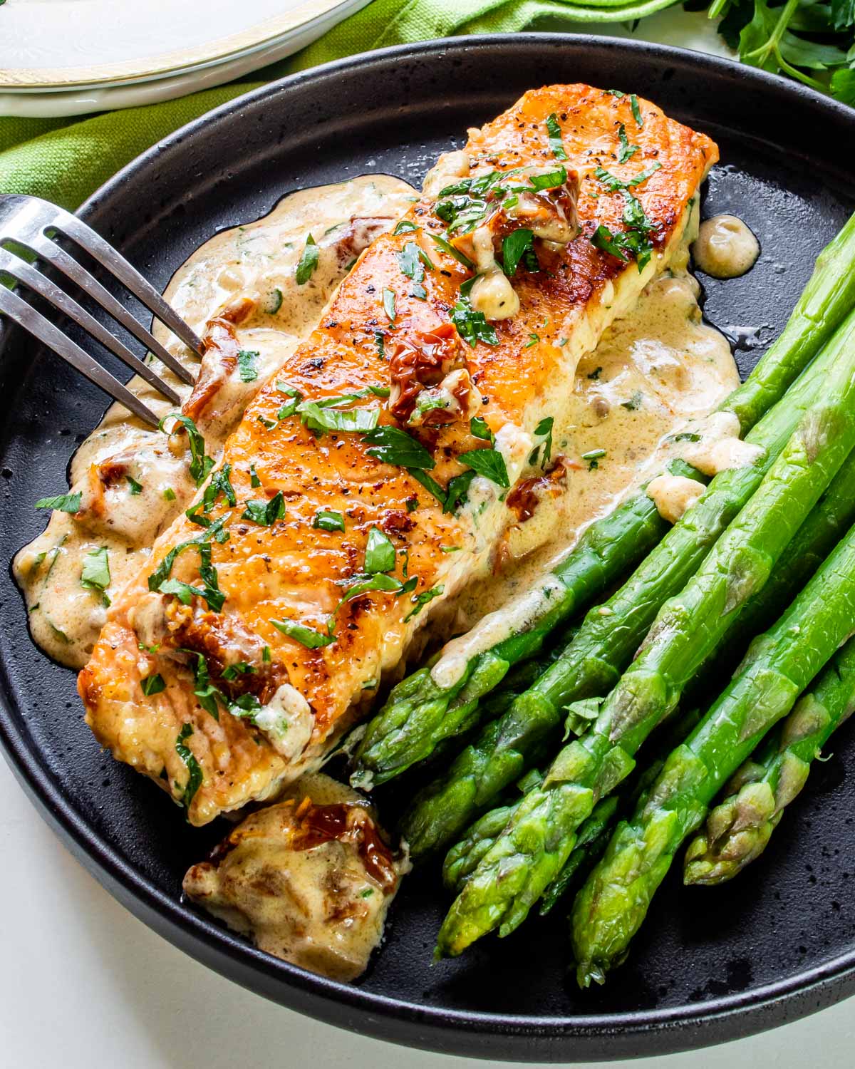 a piece of creamy tuscan salmon with some asparagus on a black plate.