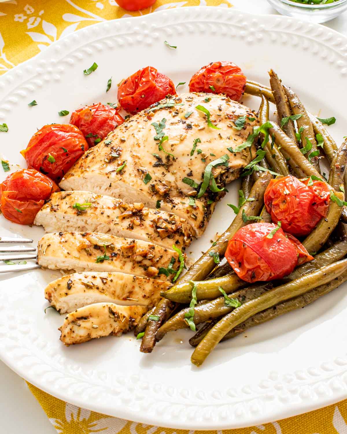 italian chicken breast sliced up with roasted green beans and tomatoes on a white plate.