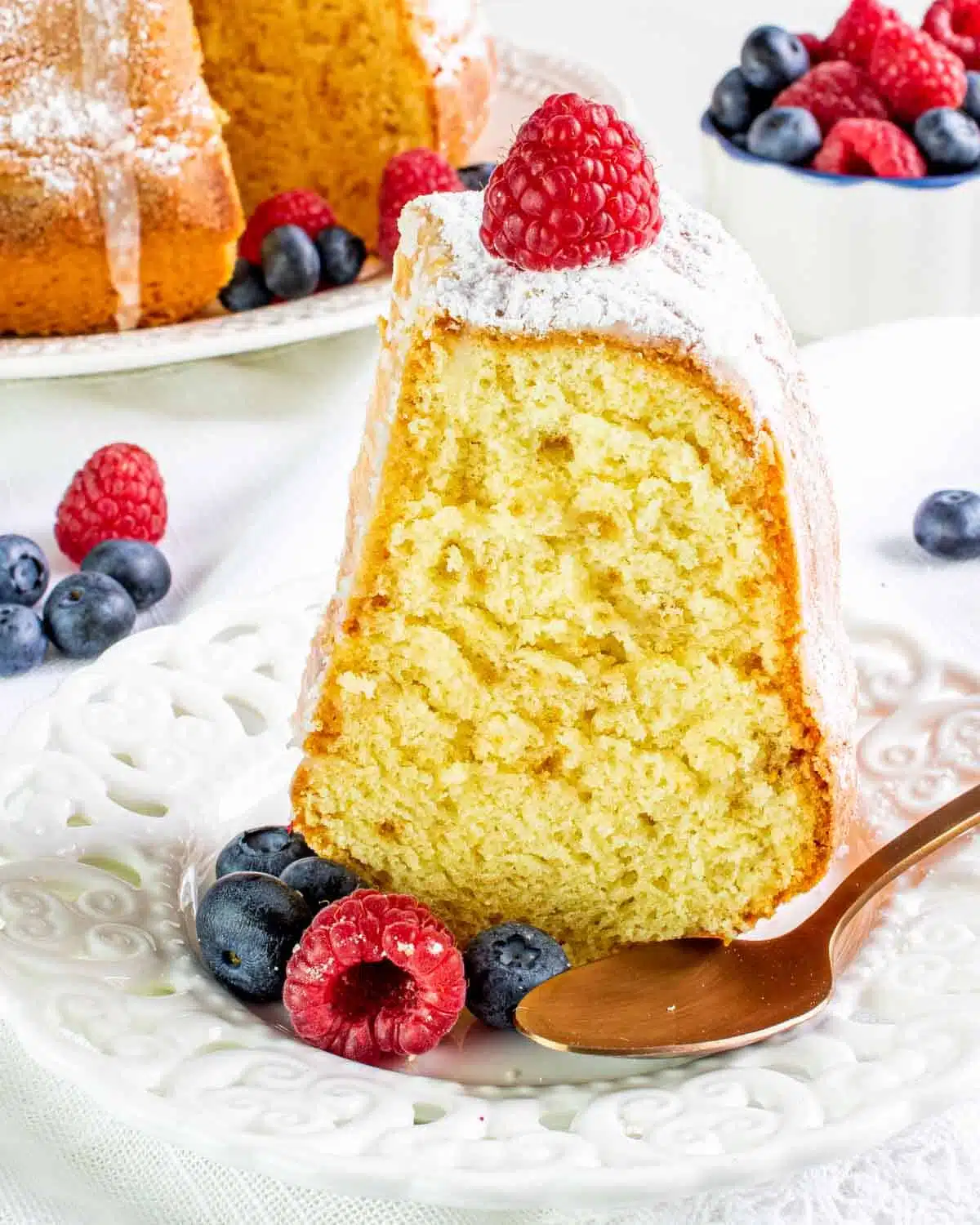 a slice of vanilla bundt cake on a dessert plate topped with berries and sprinkled with powdered sugar.