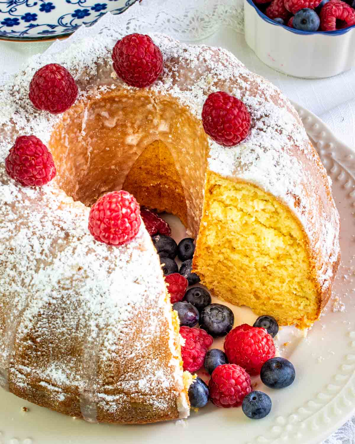a gorgeous vanilla bundt cake sprinkled with powdered sugar and topped with berries with a slice cut out of it.