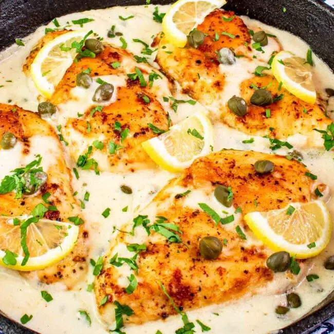 creamy chicken piccata in a black cast iron skillet garnished with parsley and lemon wedges.
