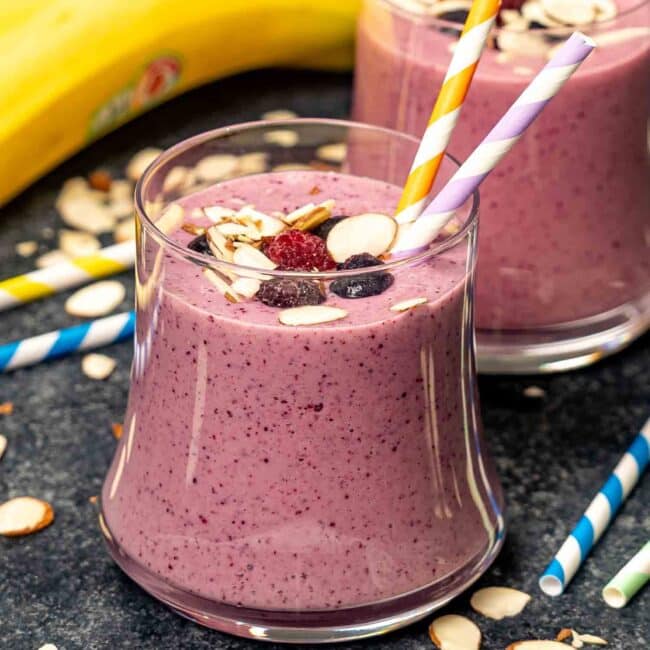 a berry and banana breakfast smoothie in a glass garnished with sliced almonds with two straws inside.