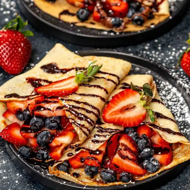two crepes stuffed with nutella and berries on a black plate.