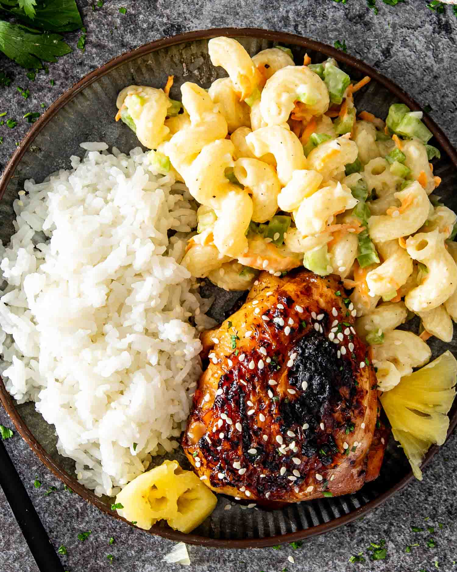 a huli huli chicken thigh on a plate along some cooked rice and macaroni salad.