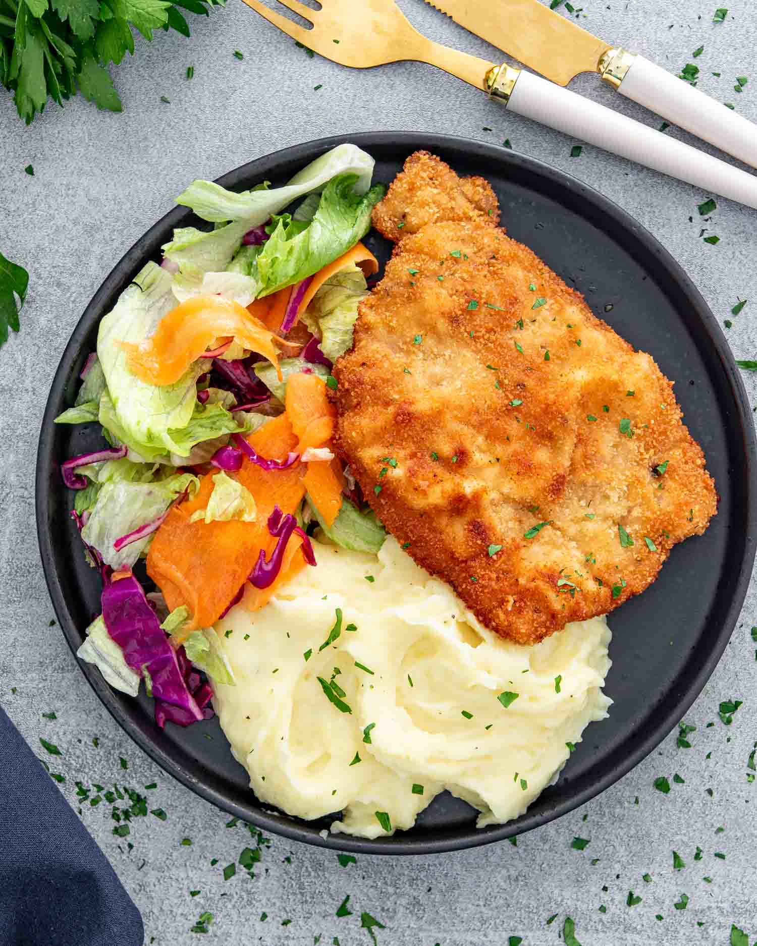 pork schnitzel with mashed potatoes and a tossed salad on a black plate.
