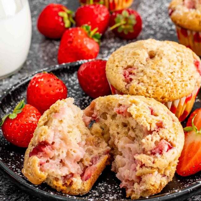 two strawberry muffins on a black plate along side some fresh strawberries.