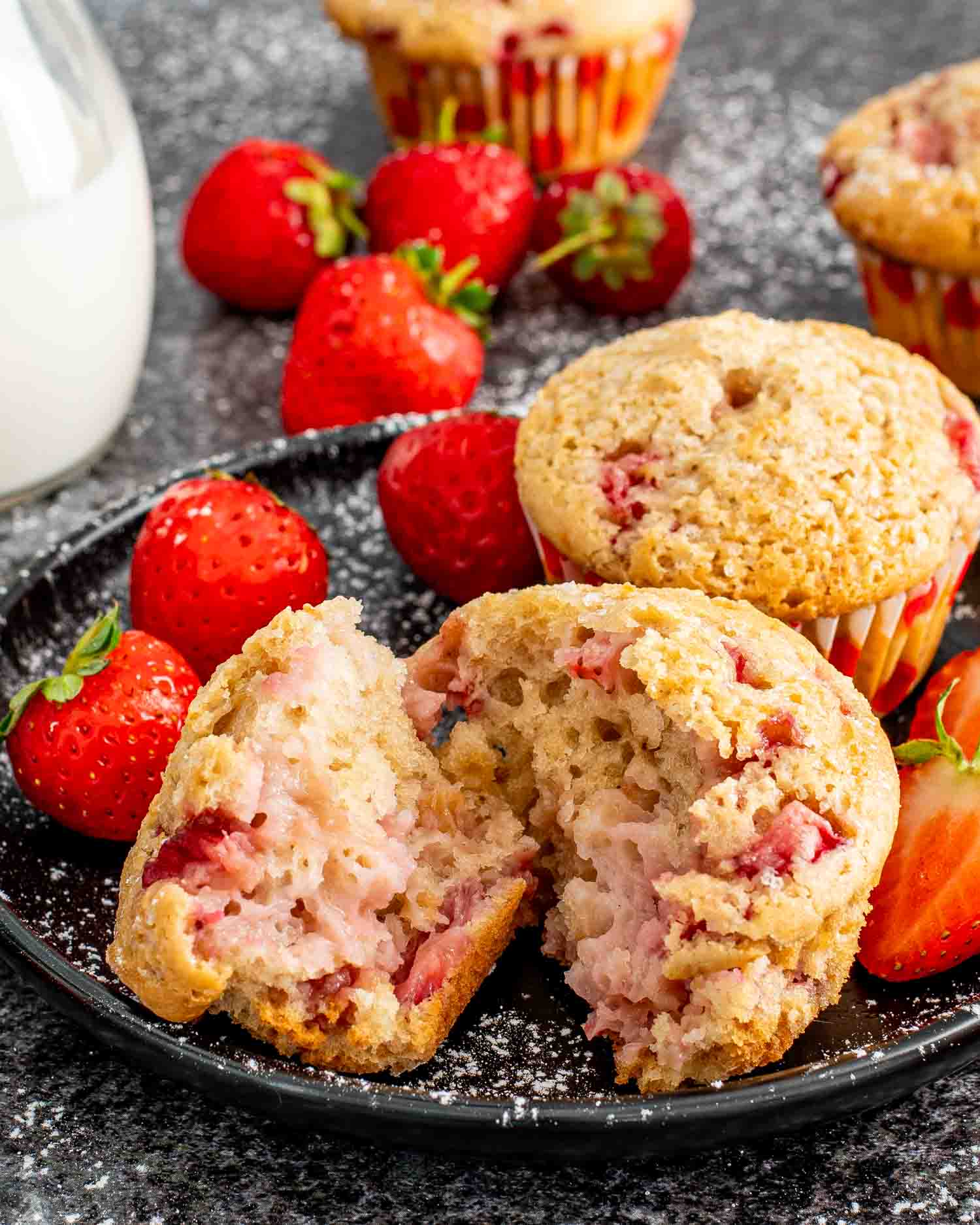 two strawberry muffins on a black plate along side some fresh strawberries.
