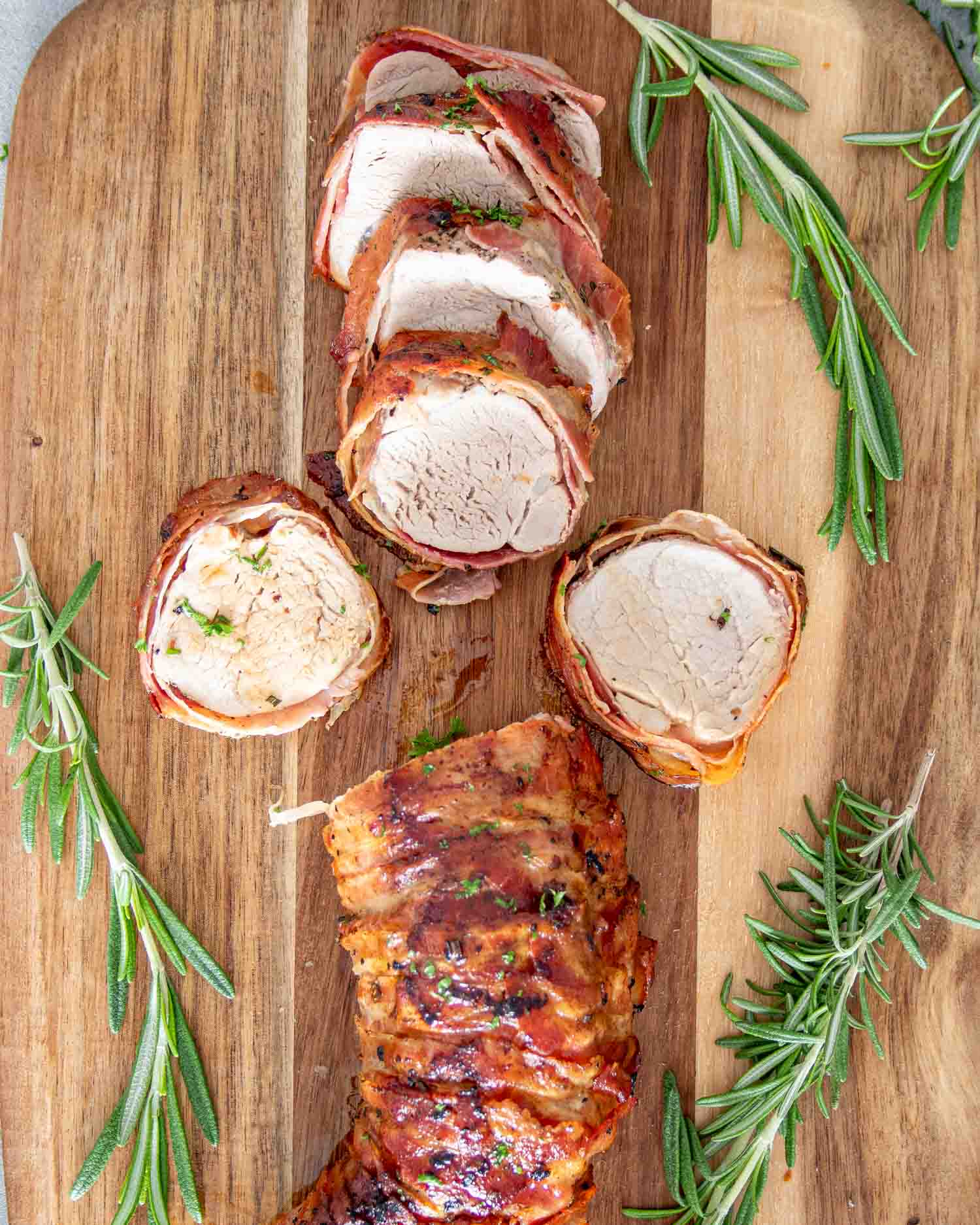 a bacon wrapped pork tenderloin cut up in slices on a cutting board along some fresh rosemary.